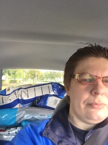 Een persoon met kort haar en een bril zit in een auto, zijn blauwe jas valt op. Op de achterbank liggen grote zakken met wat lijkt op dierenvoer, waarschijnlijk voor een donatie. Het autoraam onthult een wazig buitentafereel met groen. Een persoon met kort haar en een bril zit in een auto, zijn blauwe jas valt op. Op de achterbank liggen grote zakken met wat lijkt op dierenvoer, waarschijnlijk voor een donatie. Het autoraam onthult een wazig buitentafereel met groen.